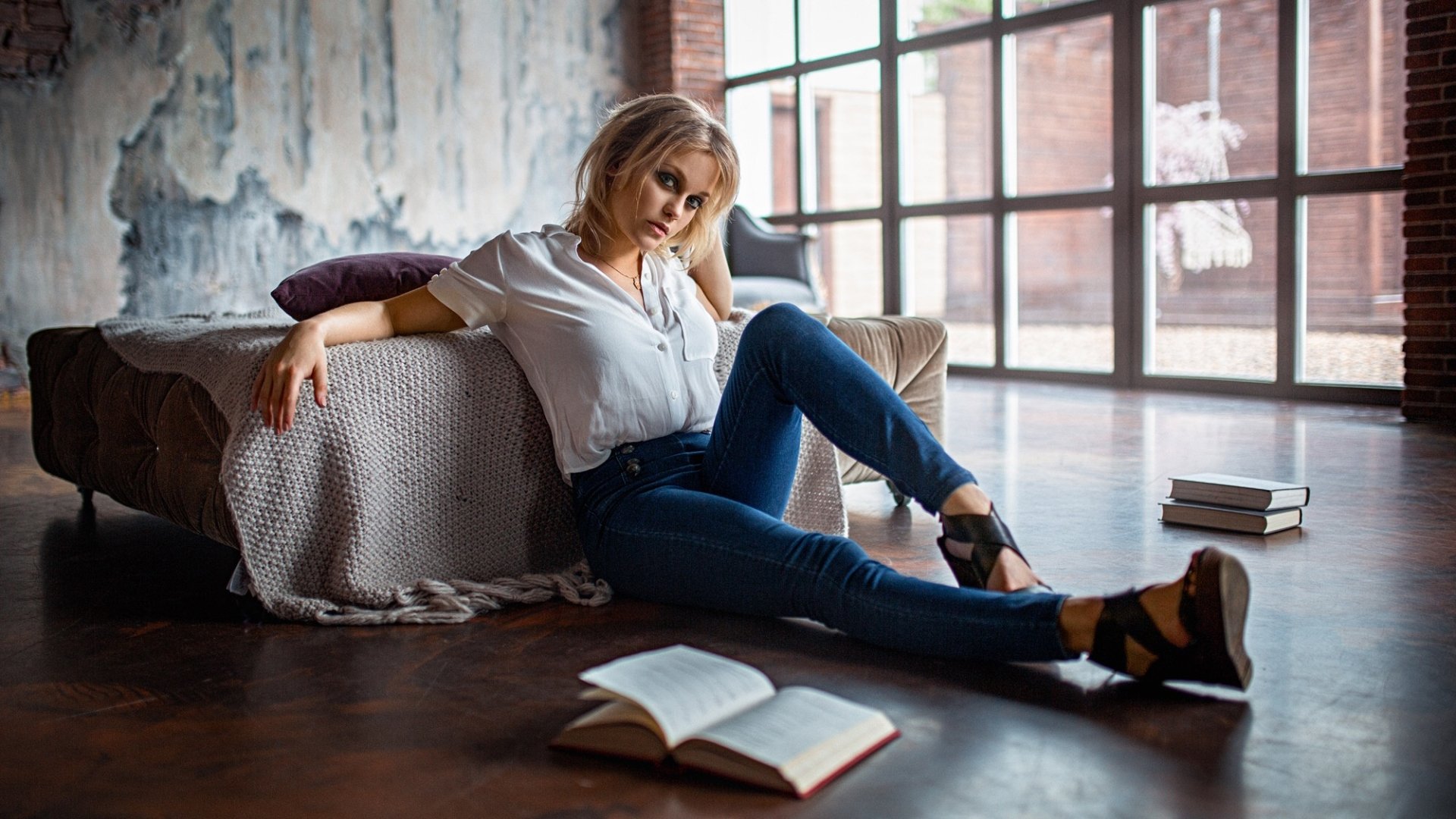 HD PC desktop wallpaper background: a pensive woman in jeans lounges on the floor by a bed, open book at her feet, moody light streaming through large industrial windows.