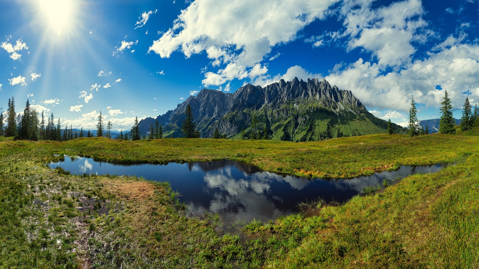 4K Ultra HD PC desktop wallpaper and background: sunbeam lights alpine meadow with spruce, pond reflecting jagged mountain and clouds under a vivid blue sky — serene nature landscape.