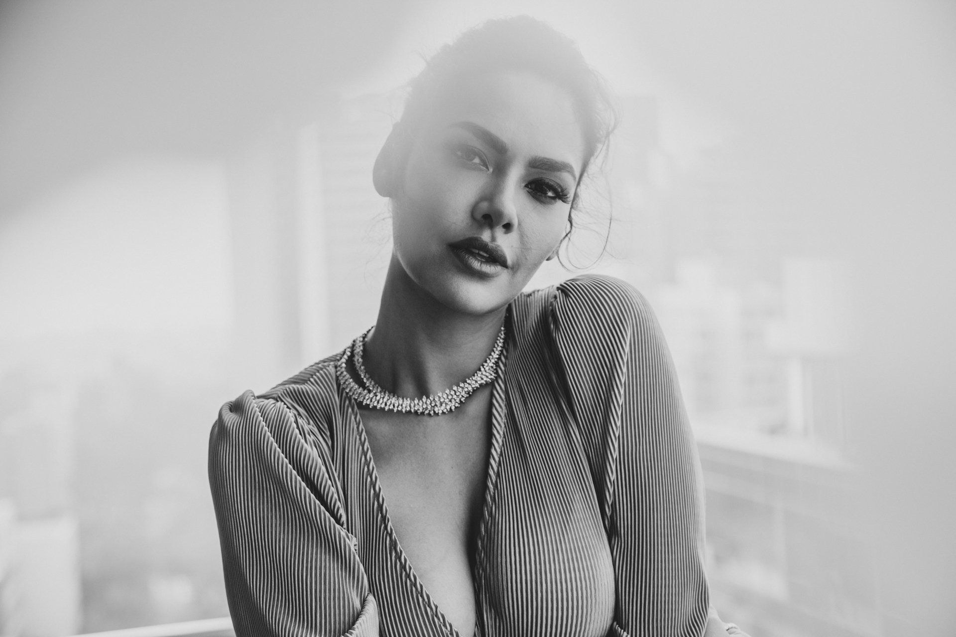 Black-and-white 2K desktop wallpaper: glamorous Bollywood-style portrait of a woman in a plunging blouse and necklace, soft city blur background, editorial model pose.