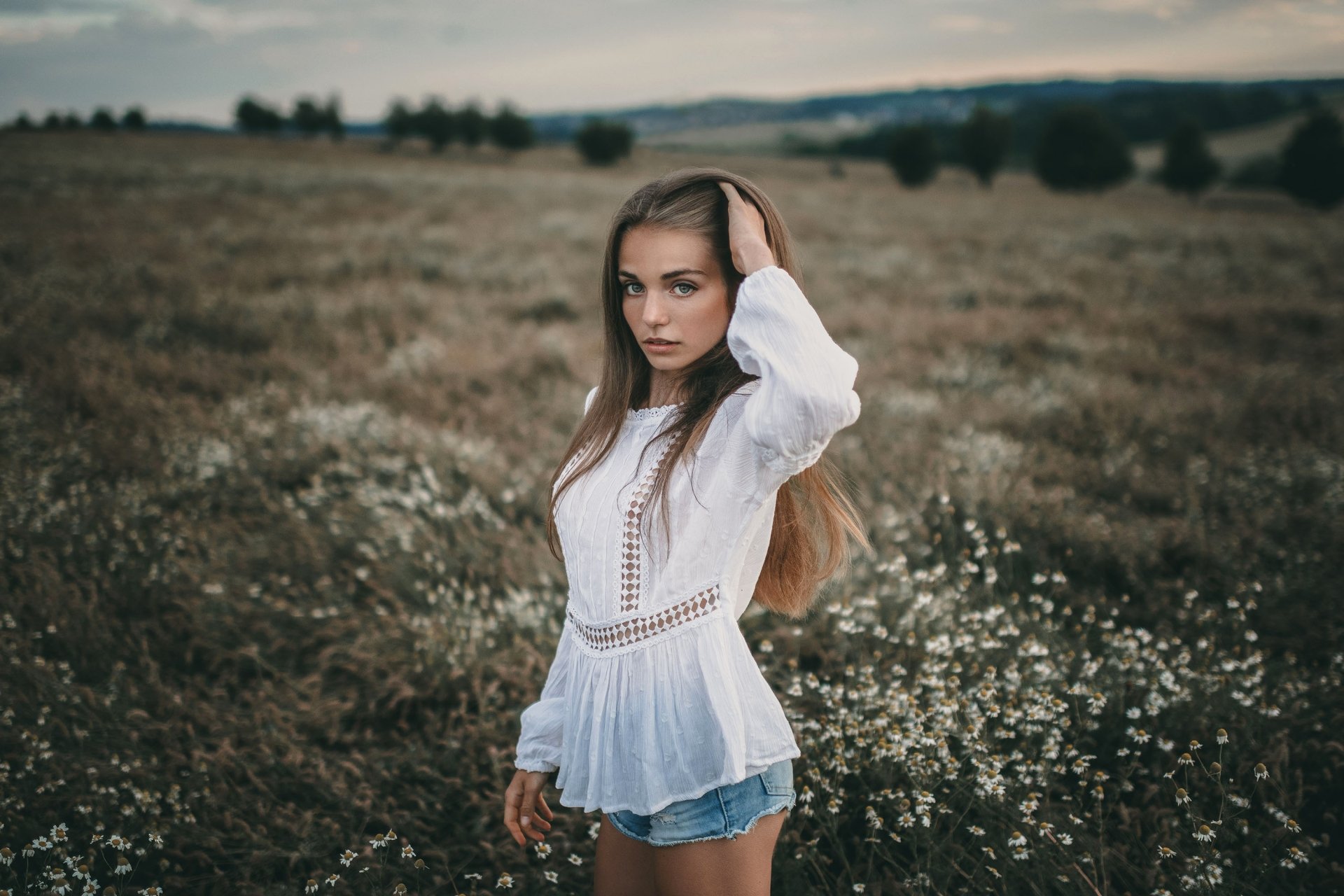 HD desktop wallpaper of a woman model with long hair wearing shorts and a white blouse, standing thoughtfully in a natural, moody outdoor setting.