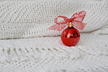 Red Christmas ornament with a sheer red bow resting on a white knitted blanket — festive holiday decoration; 5K Ultra HD PC desktop wallpaper/background.
