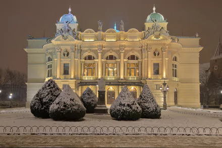 Illuminated Kraków theater building in Poland at night, surrounded by snow-covered bushes in winter, captured in HD as a desktop wallpaper background.