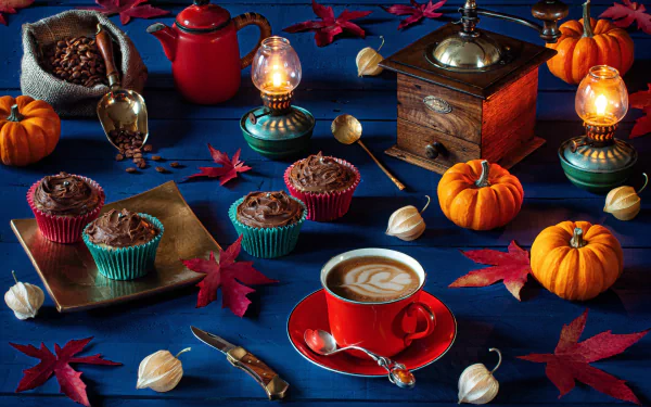 A 4K Ultra HD still life wallpaper featuring a kerosene lamp, coffee cup with latte art, cupcakes, pumpkins, a coffee grinder, and scattered maple leaves on a deep blue surface.