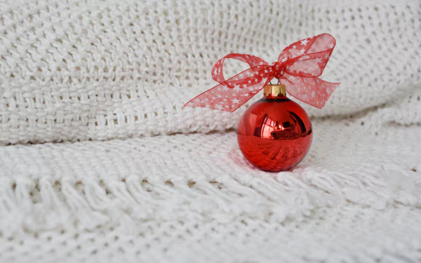 Red Christmas ornament with a sheer red bow resting on a white knitted blanket — festive holiday decoration; 5K Ultra HD PC desktop wallpaper/background.
