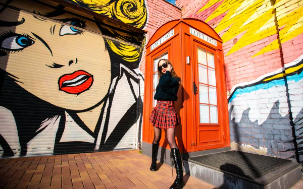 A stylish woman model wearing sunglasses, a sweater, skirt, and boots poses confidently by a red phone box against a vibrant graffiti mural, HD desktop wallpaper.