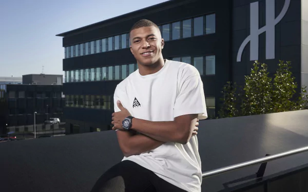 4K PC wallpaper of a smiling young French man in a white tee leaning on a rooftop railing, sports/soccer vibe with a modern building backdrop.