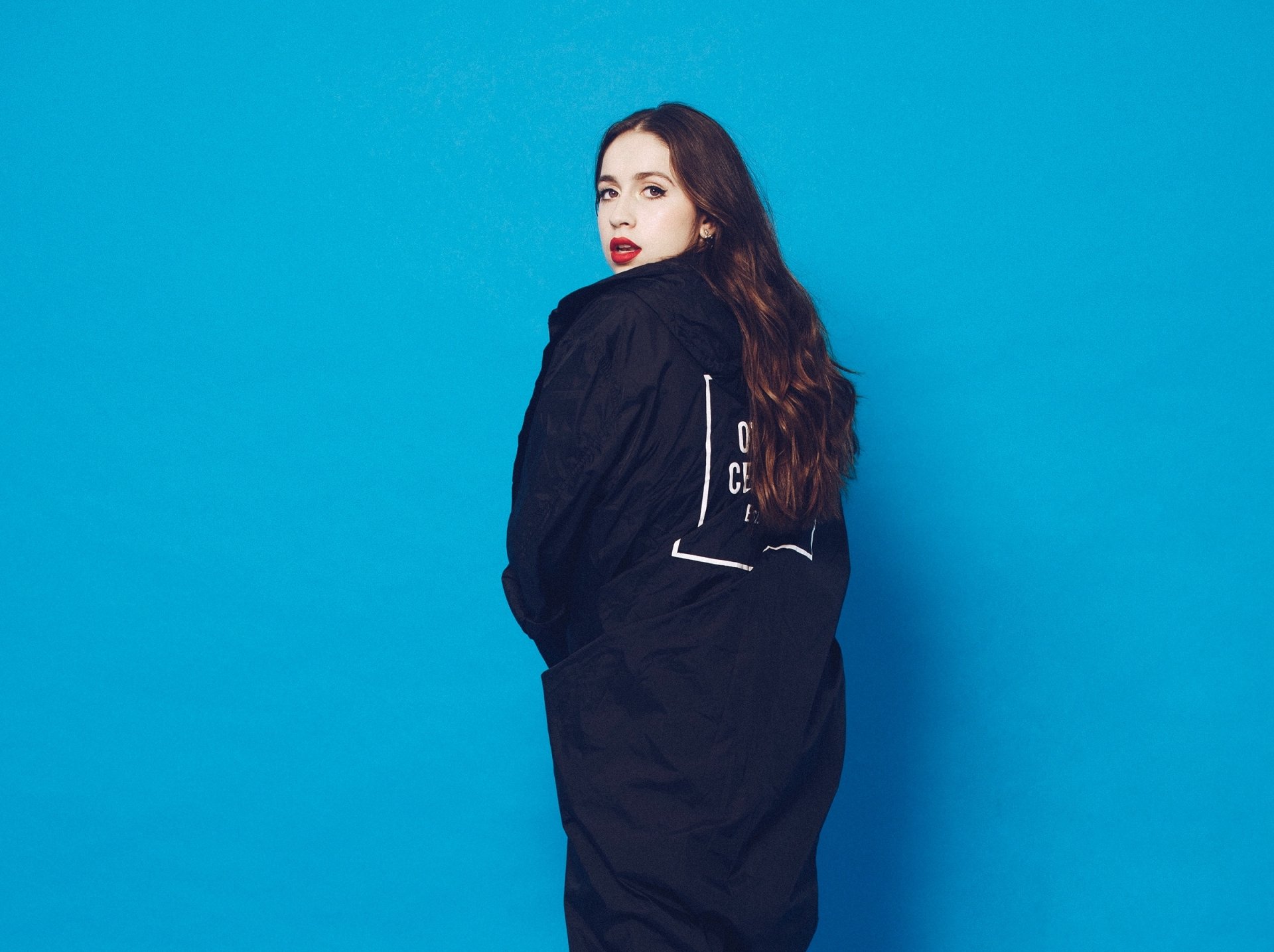 A person posing against a blue background in a black outfit, serving as a Tate McRae themed HD desktop wallpaper.