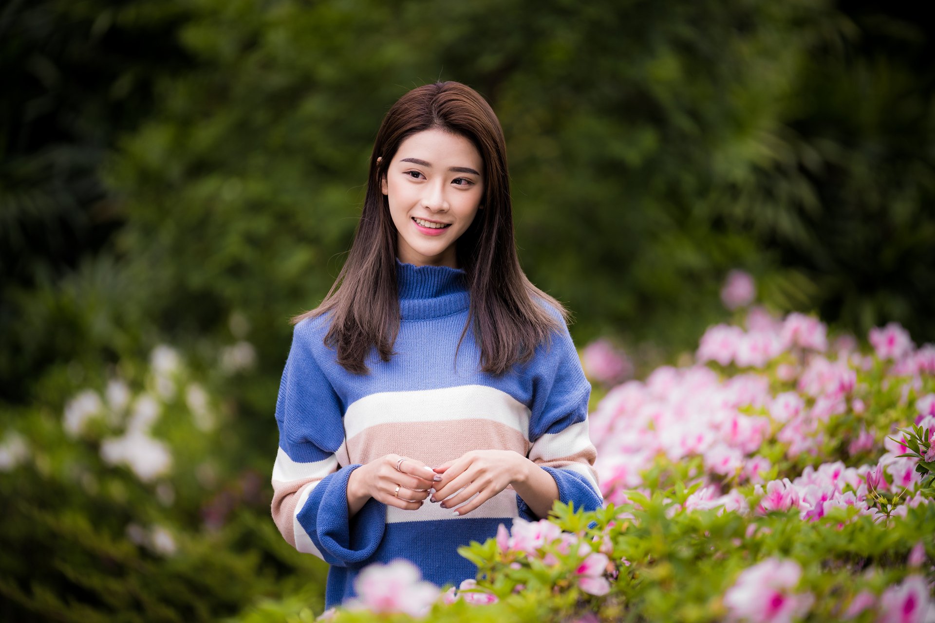 Smiling Asian brunette woman in a blue sweater stands amidst blooming flowers, captured with a depth of field effect in this 4K Ultra HD desktop wallpaper.