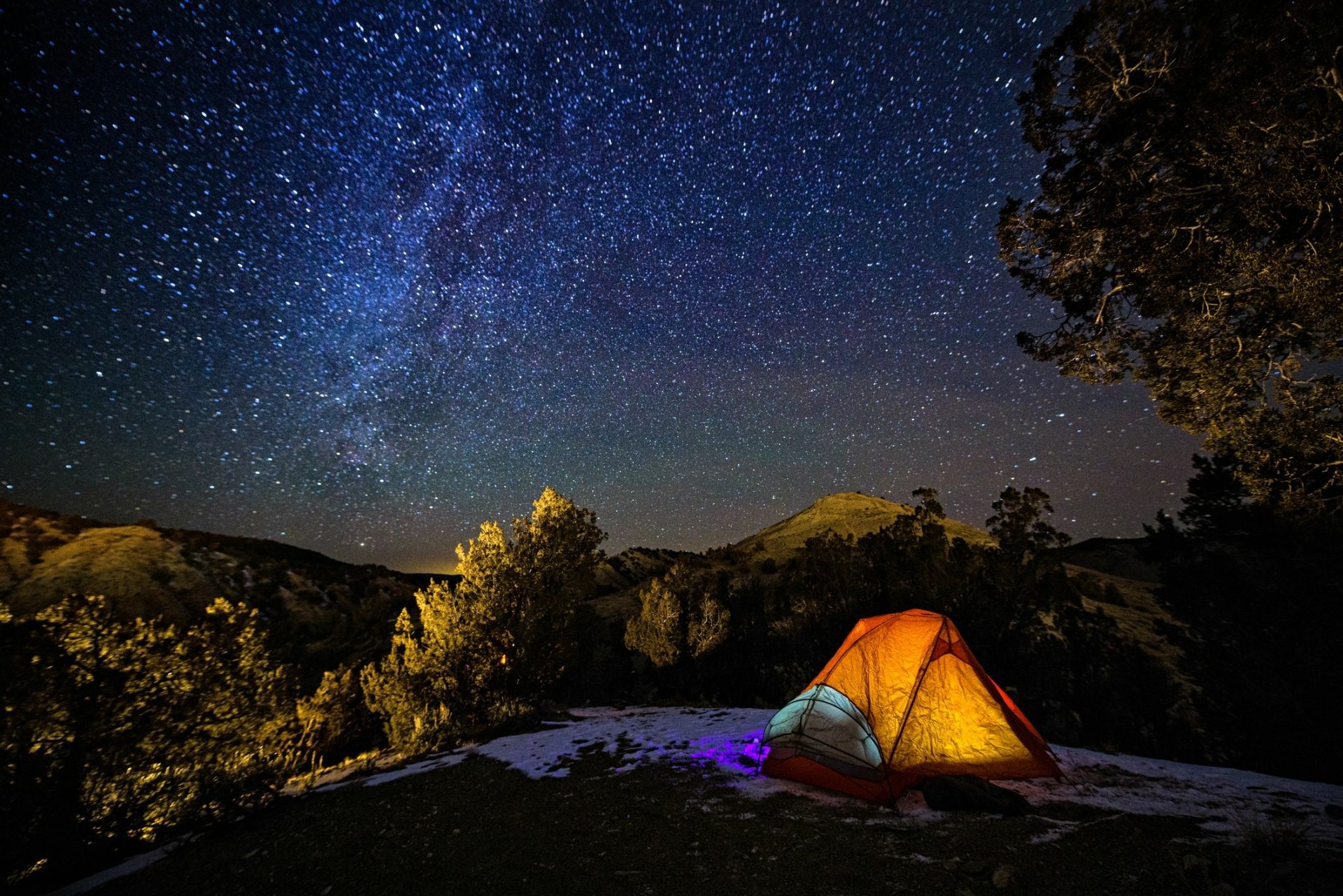 A vibrant orange tent sits under a star-filled sky, surrounded by trees and mountains, capturing the essence of camping and nature. An ideal HD desktop wallpaper.