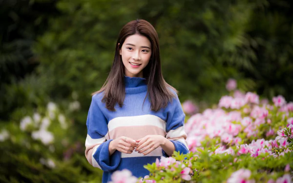 Smiling Asian brunette woman in a blue sweater stands amidst blooming flowers, captured with a depth of field effect in this 4K Ultra HD desktop wallpaper.