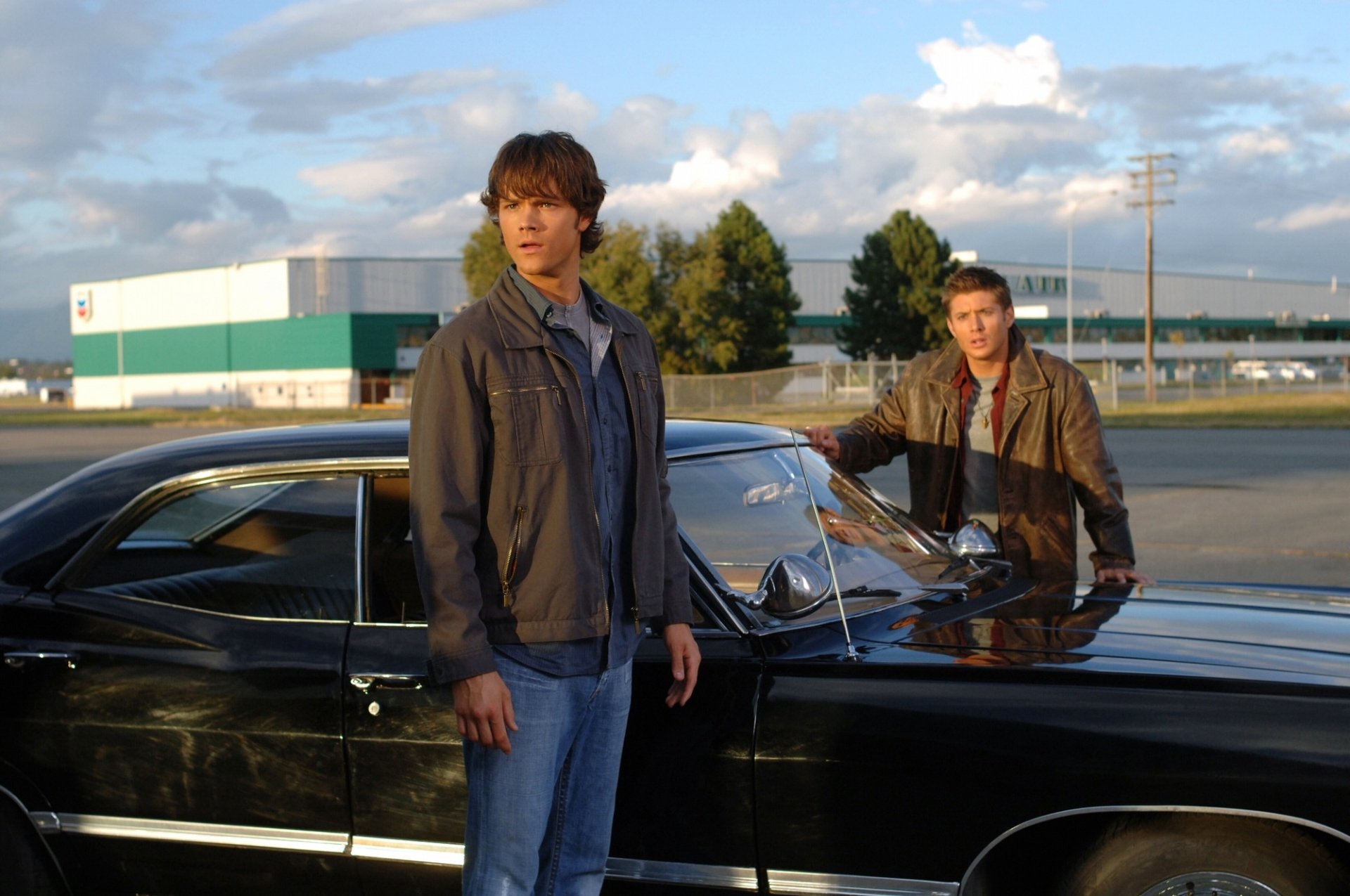 Sam Winchester and Dean Winchester from Supernatural stand by their black car in this HD PC desktop wallpaper featuring Jared Padalecki and Jensen Ackles.