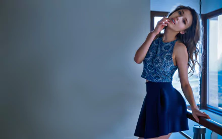 Brunette woman wearing a patterned top and navy skirt poses by a window in a bright, modern setting, captured in 4K Ultra HD for a desktop wallpaper.