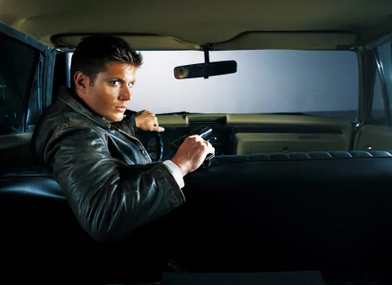 HD desktop wallpaper of Dean Winchester, portrayed by Jensen Ackles, from the TV show Supernatural, sitting in a car holding a gun.