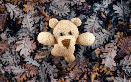 A cute, handcrafted teddy bear stands out against a backdrop of dark, textured leaves, creating a charming 4K Ultra HD desktop wallpaper and background.
