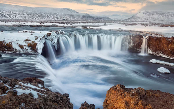 HD desktop wallpaper of Goðafoss waterfall in Iceland, showcasing cascading water amidst snowy, rugged terrain under a soft, cloudy sky.