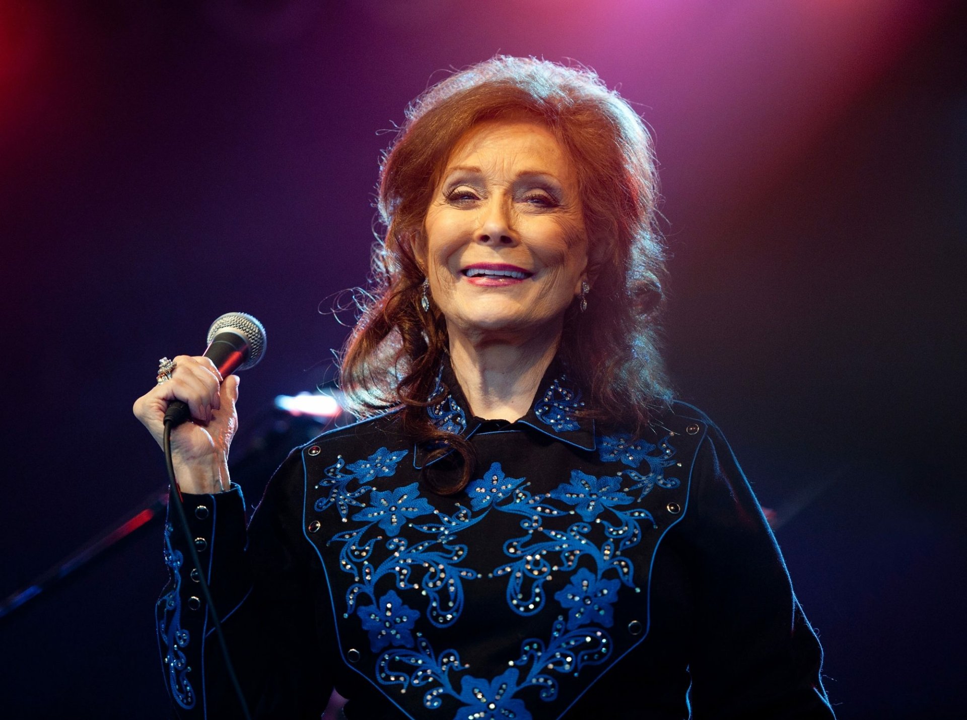 HD desktop wallpaper featuring a smiling woman with a microphone, tagged as Loretta Lynn, with a blue-lit stage background.