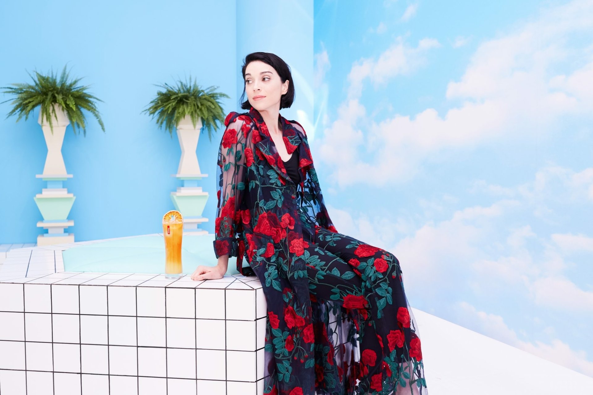 Elegant person in a floral dress sitting against a minimalist blue backdrop with decorative palms and a refreshing drink for a St. Vincent themed HD desktop wallpaper and background.