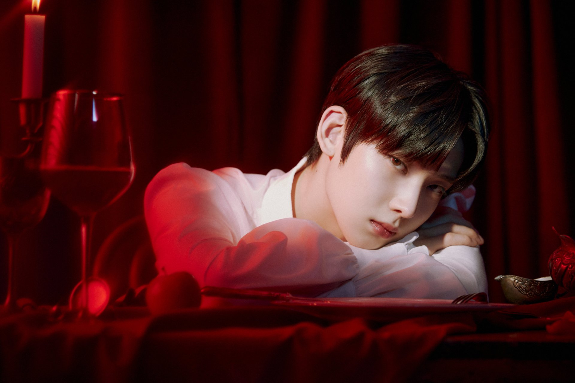 2K Quad HD music desktop wallpaper: ENHYPEN-inspired moody portrait of a pensive young man resting his head on folded arms atop a red-draped table with candles and a wine glass.