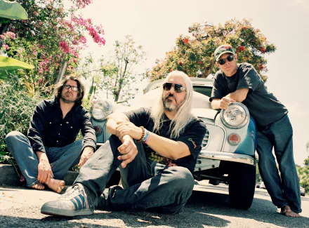 HD desktop wallpaper featuring three members of Dinosaur Jr. posing casually with a vintage car, set against lush foliage.