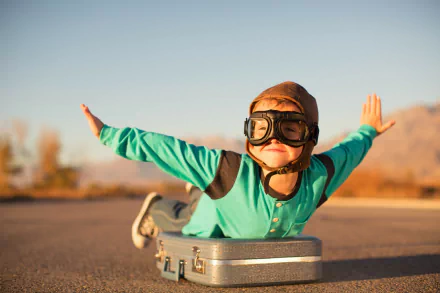 A joyful child wearing goggles and a cap lies on a suitcase, pretending to fly against a scenic backdrop, capturing the essence of imagination in this vibrant HD photography backdrop.
