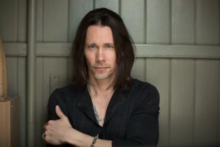Myles Kennedy HD desktop wallpaper featuring a male artist with long dark hair, wearing a black shirt, posing with crossed arms against a gray background.