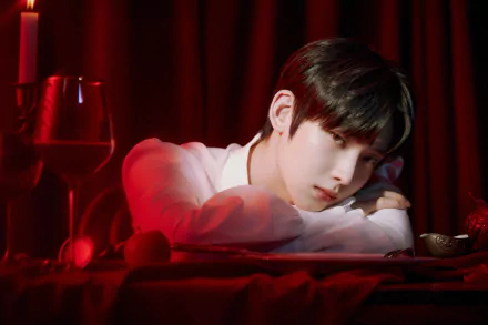 2K Quad HD music desktop wallpaper: ENHYPEN-inspired moody portrait of a pensive young man resting his head on folded arms atop a red-draped table with candles and a wine glass.