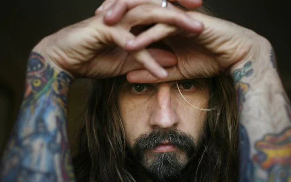 HD wallpaper of a person with long hair and full beard, looking through hands folded above eyes, with visible tattoos on arms.