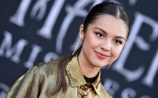 2K desktop wallpaper: close-up of a brunette American singer-actress smiling in a metallic gold outfit with sleek ponytail, photographed at a public event.