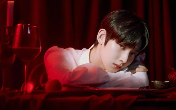 2K Quad HD music desktop wallpaper: ENHYPEN-inspired moody portrait of a pensive young man resting his head on folded arms atop a red-draped table with candles and a wine glass.