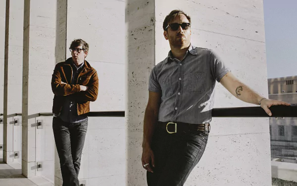 Two members of The Black Keys band posing casually, one leaning against a railing and the other standing in the background, in a high-definition desktop wallpaper.