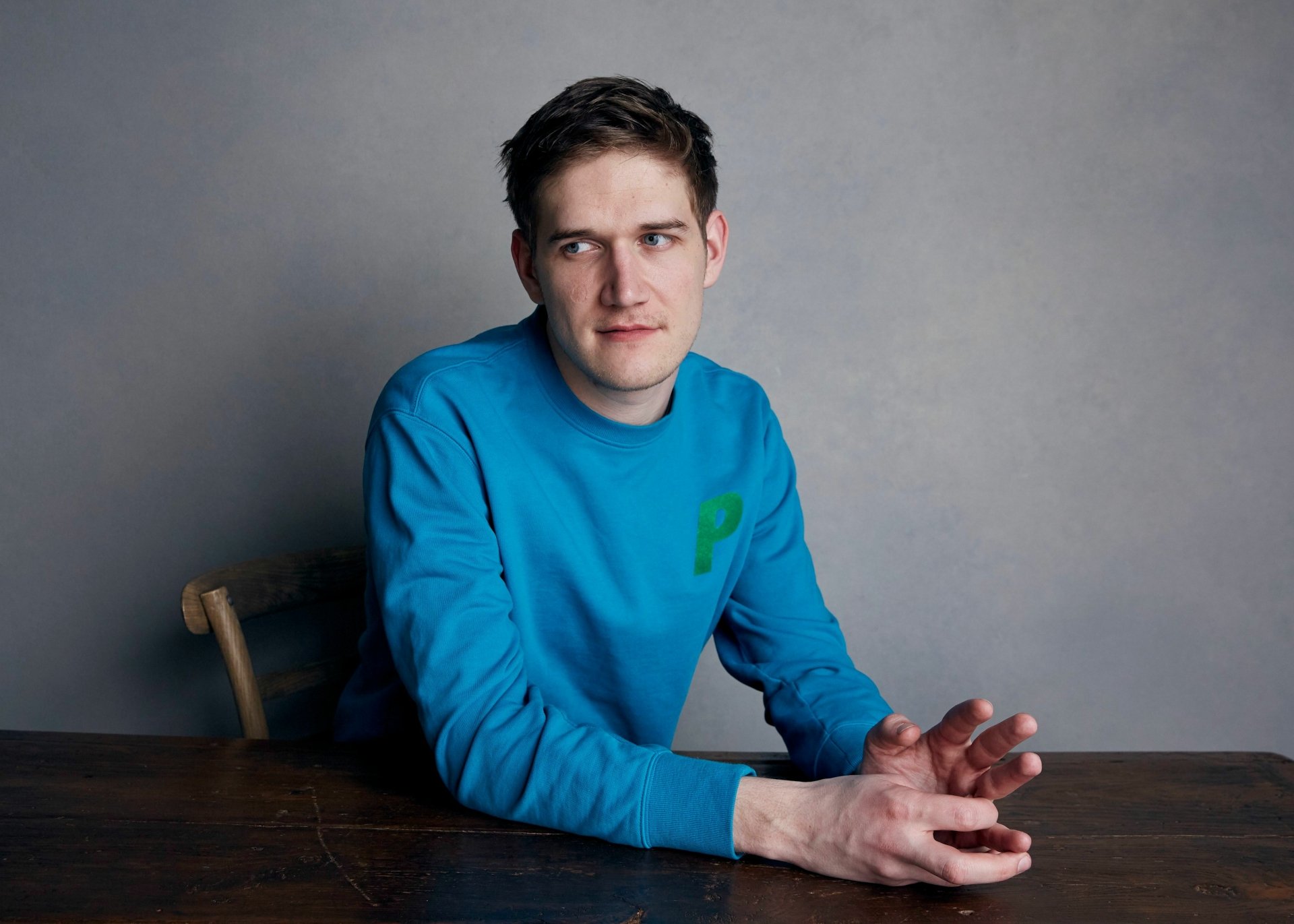 HD desktop wallpaper featuring a person seated at a table against a grey backdrop, wearing a blue sweater with a green question mark.