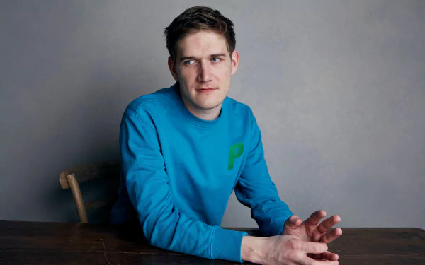 HD desktop wallpaper featuring a person seated at a table against a grey backdrop, wearing a blue sweater with a green question mark.