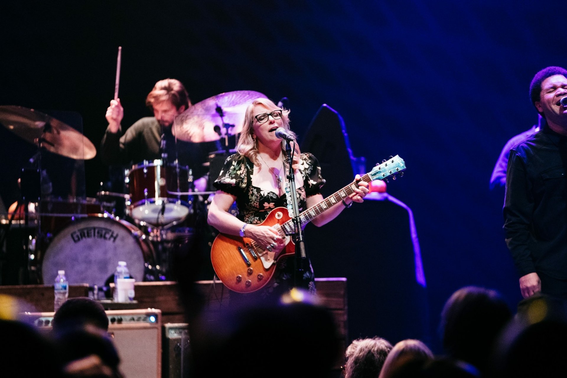 HD desktop wallpaper featuring the Tedeschi Trucks Band on stage with a guitarist in focus, a drummer in the background, and a smiling band member on the right, encapsulating the live performance energy.