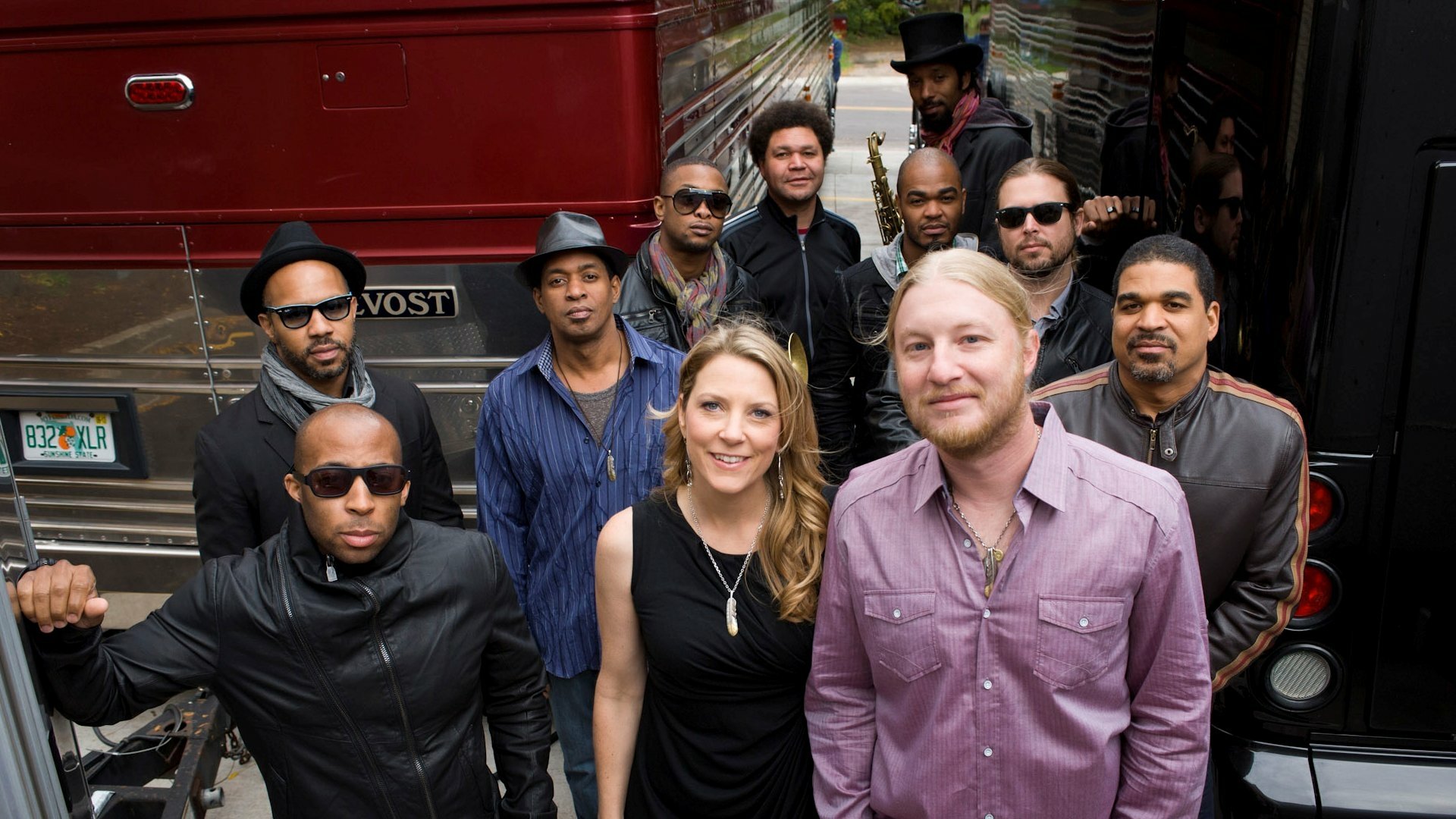 A vibrant group portrait of the Tedeschi Trucks Band, showcasing musicians in stylish outfits, standing confidently in front of a tour bus, capturing the essence of their music.