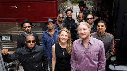 A vibrant group portrait of the Tedeschi Trucks Band, showcasing musicians in stylish outfits, standing confidently in front of a tour bus, capturing the essence of their music.