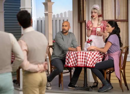 Scene from Schmigadoon! featuring two characters seated at a cafe table on a theatrical set as background for a HD desktop wallpaper.