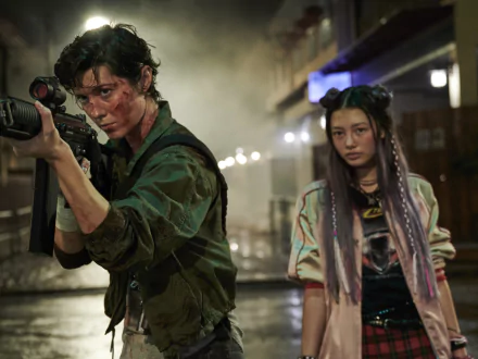 HD desktop wallpaper from the film Kate: a bloodied woman in combat gear aims a rifle while a braided‑hair teen in a colorful jacket stands beside her on a wet, neon‑lit street.