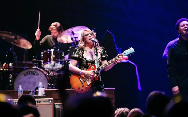 HD desktop wallpaper featuring the Tedeschi Trucks Band on stage with a guitarist in focus, a drummer in the background, and a smiling band member on the right, encapsulating the live performance energy.
