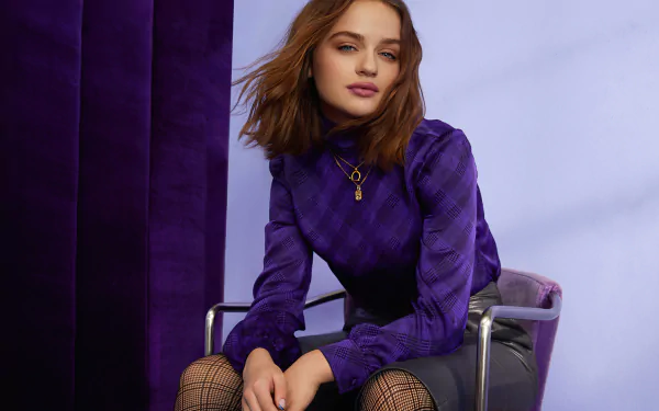 HD PC desktop wallpaper featuring American actress and celebrity Joey King seated against a purple backdrop, wearing a stylish purple top and black skirt.