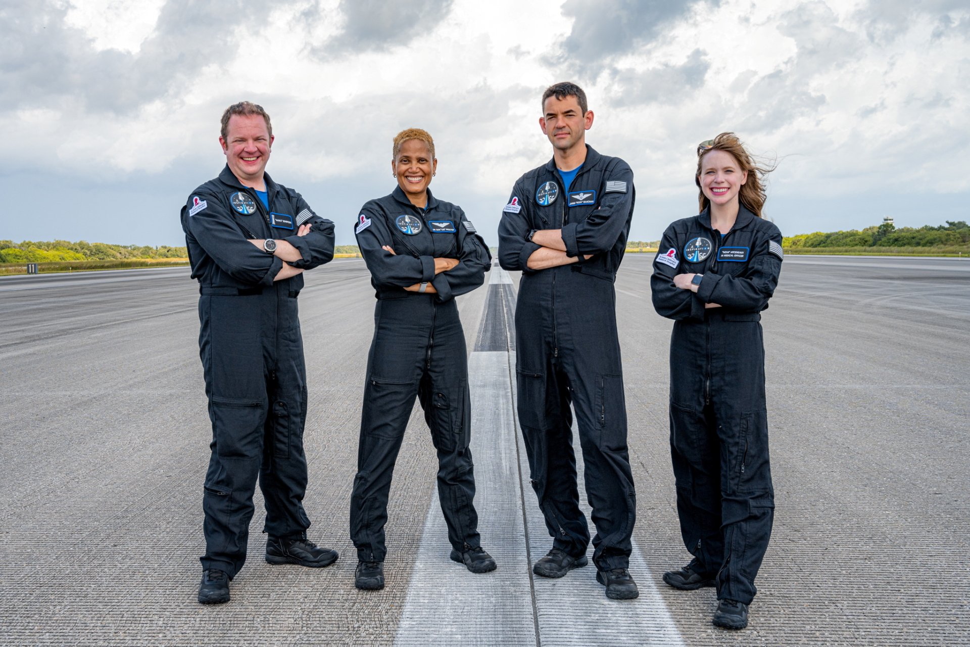 HD wallpaper featuring the Inspiration4 mission crew in flight suits, standing confidently on a runway with a cloudy sky in the background.