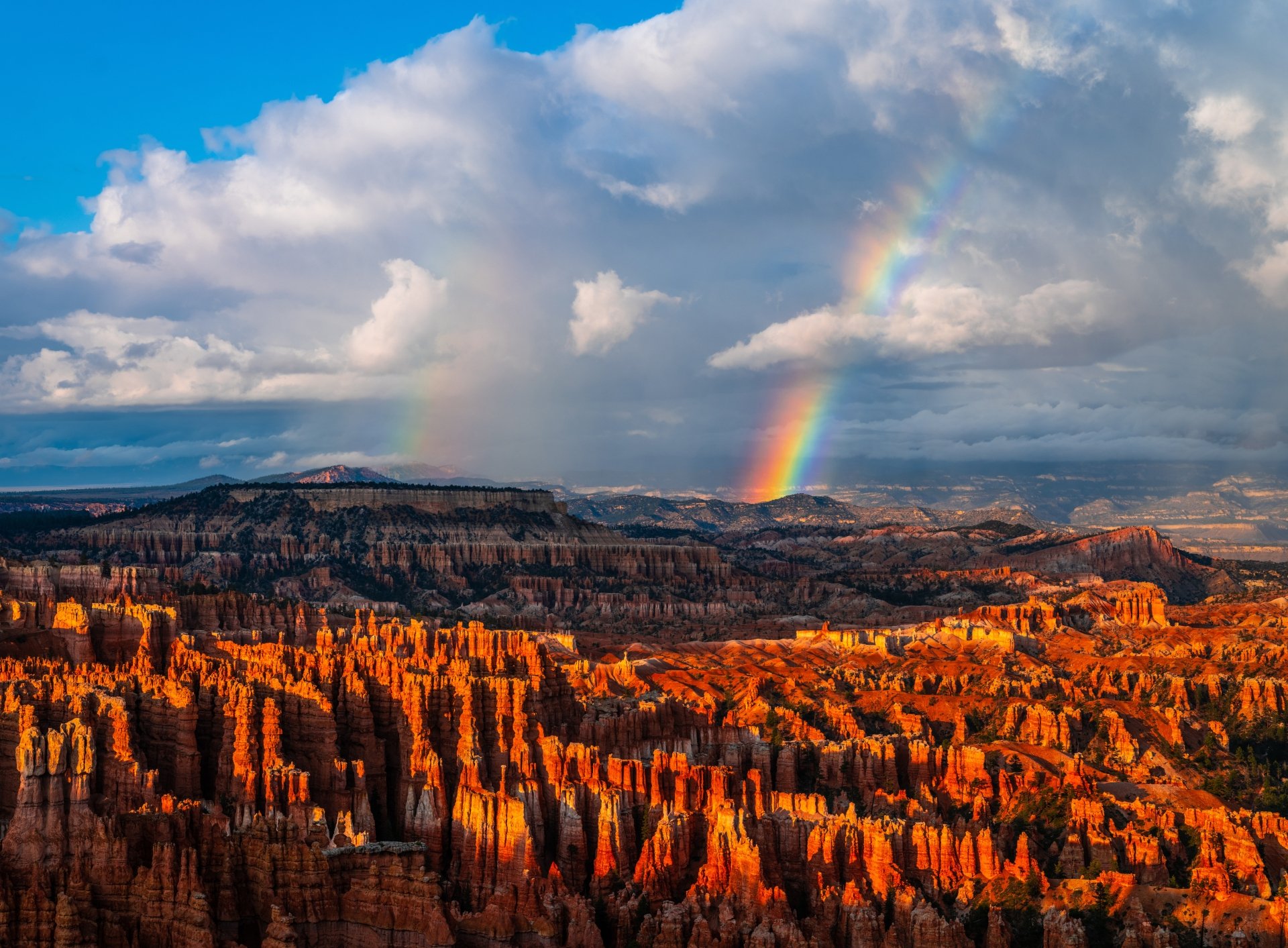 A vibrant rainbow arches over a dramatic, sunlit rocky landscape under a partly cloudy sky in this 8K Ultra HD nature desktop wallpaper.