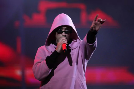 HD PC desktop wallpaper of a hooded rapper performing live, gripping a red microphone and pointing to the crowd against moody red stage lighting, conveying a music concert vibe.
