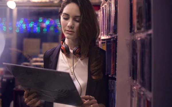 Moody 5K Ultra HD PC desktop wallpaper of a young woman with headphones holding a vinyl, leaning by shelves under soft bokeh lights.