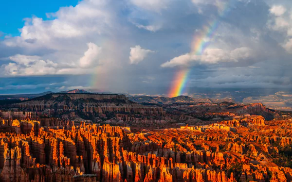 A vibrant rainbow arches over a dramatic, sunlit rocky landscape under a partly cloudy sky in this 8K Ultra HD nature desktop wallpaper.