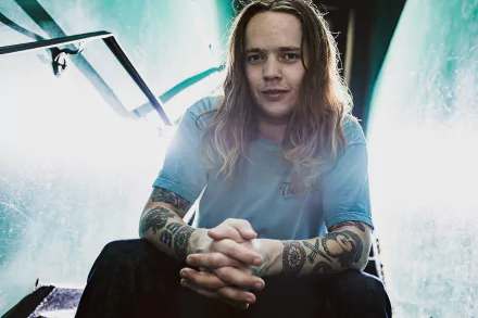 A striking HD desktop wallpaper featuring Billy Strings, showcasing his long hair and intricate tattoos in a captivating pose against a dramatic, illuminated background.