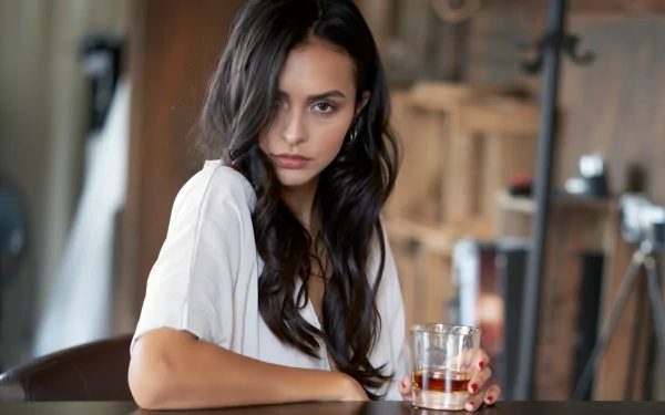 4K Ultra HD PC desktop wallpaper: black‑haired woman model with long waves, seated at a bar holding a glass, intense gaze, soft natural light and blurred background.