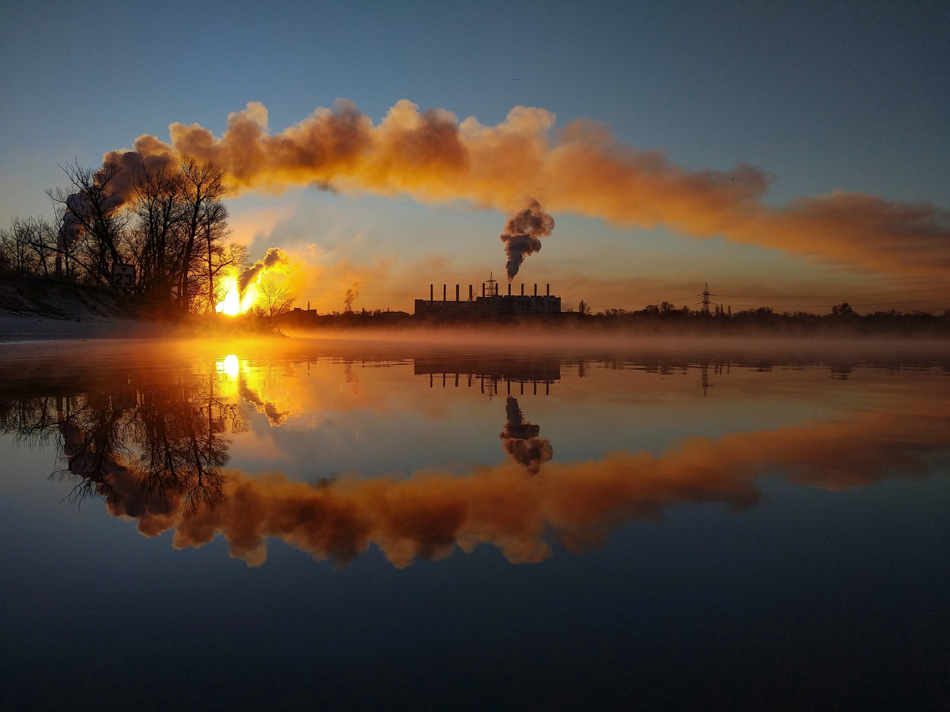 A 4K Ultra HD wallpaper showing a factory emitting smoke, reflected on a calm body of water at sunset with vibrant orange and blue hues.