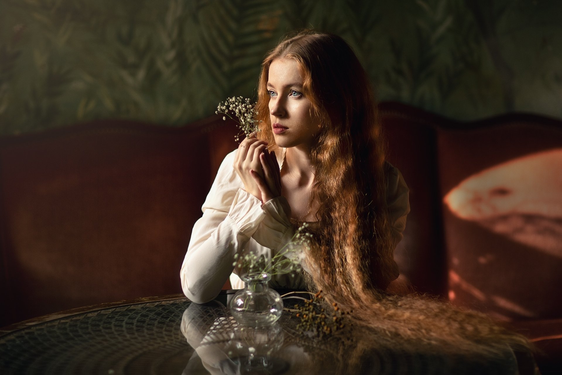 HD PC desktop wallpaper of a redhead model woman with long flowing hair in a white dress, seated at a table with small flowers, bathed in soft warm lighting.