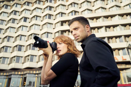 Cinematic movie still: woman aims a DSLR while a man stands beside her against a patterned urban façade — 4K Ultra HD PC desktop wallpaper and background.