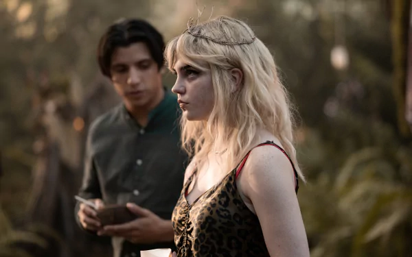 Yellowjackets TV show 4K wallpaper: pensive blonde woman with twig crown and patterned dress in a forest, a dark-haired man with a notebook stands behind.